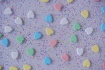 Colorful heart-shaped candies with words imprinted on them are scattered on a sparkling, confetti-style background. The candies display pastel shades of blue, green, yellow, white, and pink.