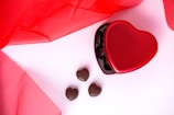 A red heart-shaped box with a shiny surface contains dark chocolate hearts. Three additional chocolate hearts are placed on a light pink surface next to the box. Sheer red fabric drapes around the composition, adding a romantic and elegant touch.