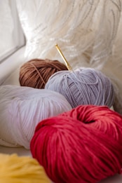 Several balls of yarn in various colors, including red, white, brown, and gray, are placed on a surface by a window. A golden crochet hook is partially inserted into the gray ball of yarn. The background features sheer, textured curtains that softly diffuse the light.