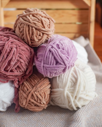 Several skeins of plush, velvet-like yarn are stacked together. The yarn is in soft, muted colors like lavender, cream, blush pink, and light brown. The setting appears cozy, with the yarns placed on a woven fabric surface and wooden background, suggesting a crafting or knitting environment.