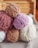 Several skeins of plush, velvet-like yarn are stacked together. The yarn is in soft, muted colors like lavender, cream, blush pink, and light brown. The setting appears cozy, with the yarns placed on a woven fabric surface and wooden background, suggesting a crafting or knitting environment.