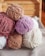 Several skeins of plush, velvet-like yarn are stacked together. The yarn is in soft, muted colors like lavender, cream, blush pink, and light brown. The setting appears cozy, with the yarns placed on a woven fabric surface and wooden background, suggesting a crafting or knitting environment.