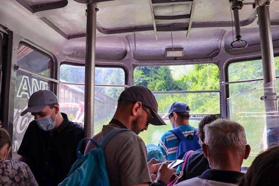 A group of people inside a cable car with windows showing a lush green landscape outside. Some individuals are wearing caps and backpacks, and one person is looking at their phone. The setting appears to be a casual travel or tourism environment.