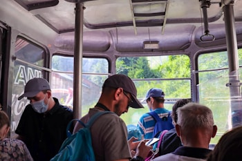 A group of people inside a cable car with windows showing a lush green landscape outside. Some individuals are wearing caps and backpacks, and one person is looking at their phone. The setting appears to be a casual travel or tourism environment.