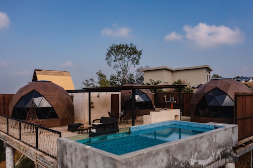Geodesic dome structures are situated on a terrace, alongside a modern outdoor swimming pool. The setting includes outdoor furniture like chairs and tables, with trees and clear blue sky in the background.