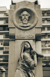 A stone monument featuring a carved portrait in a circular frame at the top, labeled Avellaneda. Below, a sculpted figure of a woman in robes is depicted in a pleading pose, looking upward. The monument is set against a blurred background of modern buildings.