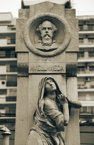 A stone monument featuring a carved portrait in a circular frame at the top, labeled Avellaneda. Below, a sculpted figure of a woman in robes is depicted in a pleading pose, looking upward. The monument is set against a blurred background of modern buildings.