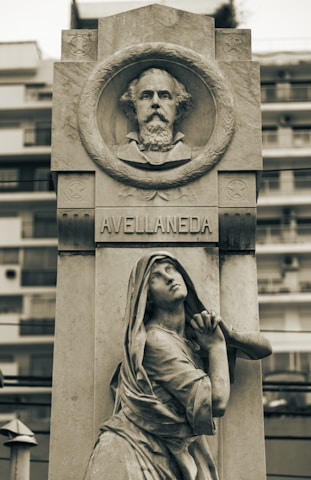 A stone monument featuring a carved portrait in a circular frame at the top, labeled Avellaneda. Below, a sculpted figure of a woman in robes is depicted in a pleading pose, looking upward. The monument is set against a blurred background of modern buildings.