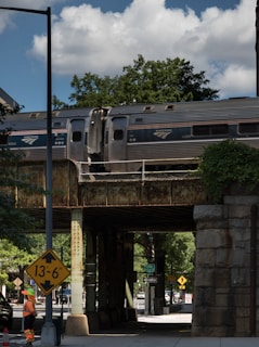 A passenger train is moving on an elevated railway bridge above a street. Below the bridge, a construction worker wearing an orange safety vest and helmet stands near a height restriction sign indicating a clearance of 13 feet 6 inches. The scene is set under a partly cloudy sky with trees and urban infrastructure visible in the background.