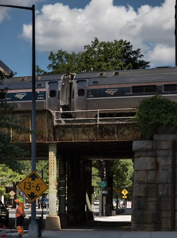 A passenger train is moving on an elevated railway bridge above a street. Below the bridge, a construction worker wearing an orange safety vest and helmet stands near a height restriction sign indicating a clearance of 13 feet 6 inches. The scene is set under a partly cloudy sky with trees and urban infrastructure visible in the background.
