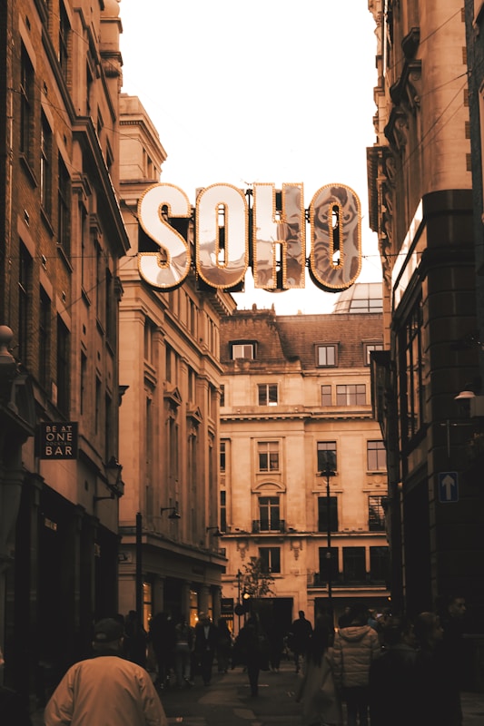 A city street scene with the focus on large, illuminated marquee letters spelling 'SOHO' suspended between buildings. The street is flanked by tall, classical-style buildings with large windows. There is a crowd of people walking beneath, and a sign for 'Be At One Bar' is visible on the left. The photo has a warm, sepia tone.
