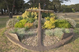 A large, weathered anchor is set prominently within a landscaped garden area, surrounded by various lush plants and flowers. The background reveals a park-like setting with tall trees and open grassy areas.