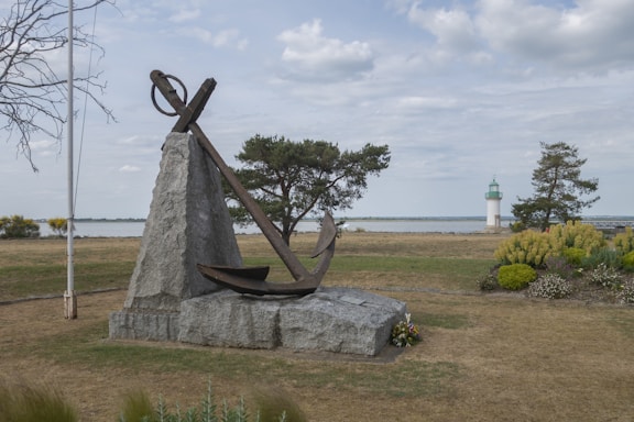 A large metal anchor mounted on a stone platform with a scenic background. The area is landscaped with various trees and a lighthouse visible in the distance by the sea. The sky is partly cloudy, and the surroundings are grassy with some bushes and flowers.