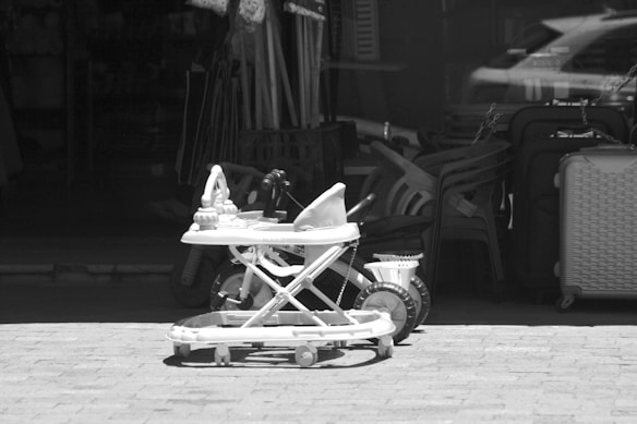 A black and white image featuring a baby walker with wheels in front of a shop. The shop displays a variety of items, including stacked plastic chairs and a suitcase. The scene captures a somewhat cluttered assortment of everyday objects.