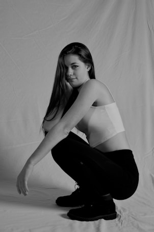 A black and white image of a woman squatting. She has long hair and is wearing a sleeveless top with leggings and boots. The background appears to be a plain fabric backdrop, creating a simple and clean aesthetic.