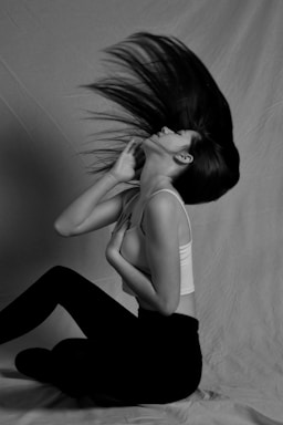 A woman is captured in mid-motion, flipping her long hair dramatically. She is wearing a white tank top and black leggings, seated on the ground against a plain backdrop. Her posture and the movement of her hair suggest a sense of freedom and dynamic energy.