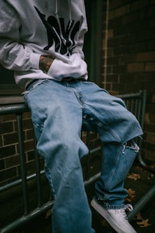 a man sitting on a rail wearing a white hoodie and blue jeans