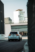 A white SUV is driving down a quiet urban street with a large building in the background featuring signage for 'Storage' and 'Green Box'. The scene has a modern, industrial feel with sleek architecture and muted tones.