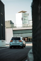 A white SUV is driving down a quiet urban street with a large building in the background featuring signage for 'Storage' and 'Green Box'. The scene has a modern, industrial feel with sleek architecture and muted tones.