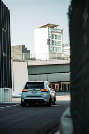 A white SUV is driving down a quiet urban street with a large building in the background featuring signage for 'Storage' and 'Green Box'. The scene has a modern, industrial feel with sleek architecture and muted tones.