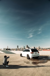 A person crouches on a rooftop parking area, photographing a white SUV with a city skyline in the background. The sky is clear with a few clouds, and the vehicle casts a shadow on the concrete. The scene conveys an urban atmosphere with a focus on the car and its surroundings.