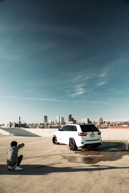 A person crouches on a rooftop parking area, photographing a white SUV with a city skyline in the background. The sky is clear with a few clouds, and the vehicle casts a shadow on the concrete. The scene conveys an urban atmosphere with a focus on the car and its surroundings.