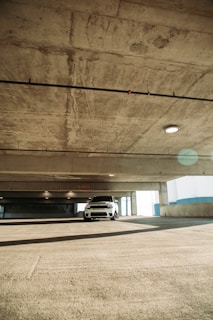 A white car is parked in a large, empty concrete parking garage. The garage is lit by natural light streaming through large openings, and the concrete surfaces show signs of wear and textures. The ceiling has exposed pipes and fixtures, and there is a speed limit sign indicating 10 MPH.