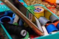 A sewing kit with various spools of thread in diverse colors including yellow, blue, green, black, and orange. A smartphone rests on the edge of the kit, partially visible. The threads and other sewing tools are organized inside a turquoise container on a patterned surface.