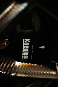 A close-up view of a carbon fiber texture, highlighting a component labeled with the word 'Koenigsegg'. The carbon fiber patterns appear shiny and reflect light, giving a glossy effect to the surface.