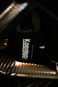 A close-up view of a carbon fiber texture, highlighting a component labeled with the word 'Koenigsegg'. The carbon fiber patterns appear shiny and reflect light, giving a glossy effect to the surface.