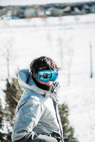 A person is wearing a winter jacket with a high collar and reflective ski goggles. The background is a snowy landscape with some trees visible, suggesting a winter setting.