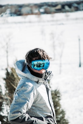 A person wearing a winter jacket and blue reflective ski goggles stands outdoors in a snowy landscape. Trees and a blurred cityscape are visible in the background, with the reflection in the goggles showing other skiers or snowboarders.