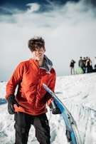 A person wearing a bright red jacket stands on a snowy landscape holding a snowboard. The foreground shows clear details of snow and the snowboard. In the background, a group of people dressed in winter clothing can be seen, with a cloudy sky above.