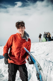 A person wearing a bright red jacket stands on a snowy landscape holding a snowboard. The foreground shows clear details of snow and the snowboard. In the background, a group of people dressed in winter clothing can be seen, with a cloudy sky above.
