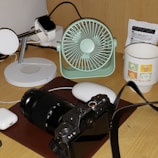 A desktop setup featuring a digital camera with a zoom lens placed on a brown mat. Nearby, a mint green portable fan and a black smartwatch on a white charging stand are visible. A white mouse and a pair of wireless earphones are also present, along with a white mug with colorful graphics and text. A small packet, possibly containing instructions or information, is located at the back.