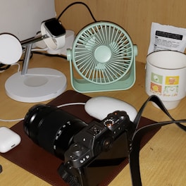 A desktop setup featuring a digital camera with a zoom lens placed on a brown mat. Nearby, a mint green portable fan and a black smartwatch on a white charging stand are visible. A white mouse and a pair of wireless earphones are also present, along with a white mug with colorful graphics and text. A small packet, possibly containing instructions or information, is located at the back.