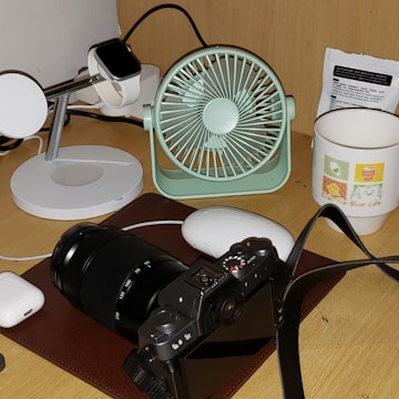 A desktop setup featuring a digital camera with a zoom lens placed on a brown mat. Nearby, a mint green portable fan and a black smartwatch on a white charging stand are visible. A white mouse and a pair of wireless earphones are also present, along with a white mug with colorful graphics and text. A small packet, possibly containing instructions or information, is located at the back.