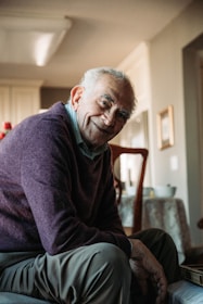 An elderly man with a gentle smile, wearing a purple sweater and sitting in a warm, well-lit room with a soft ambiance. The background suggests a cozy domestic setting with a table and kitchen visible.