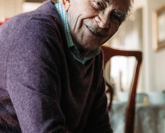 A friendly support worker tidying a sunlit living room while an elderly man relaxes nearby.