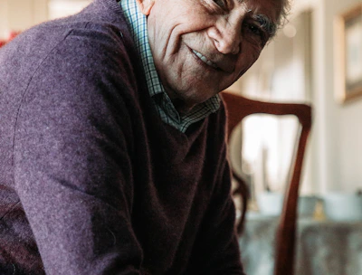 A friendly support worker tidying a sunlit living room while an elderly man relaxes nearby.
