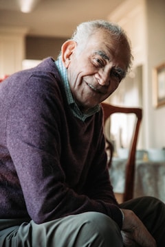 An elderly man is sitting in a warmly lit room, wearing a dark purple sweater over a checkered shirt. He is smiling gently, conveying a friendly and approachable demeanor. The background is softly focused, with hints of furniture and decor.