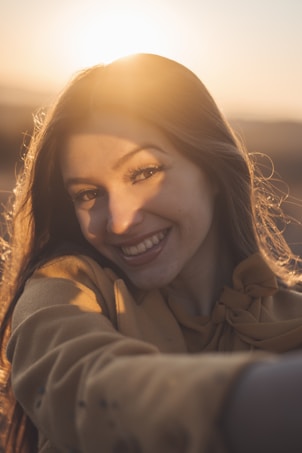A person smiling brightly with the sun setting in the background, creating a warm and soft glow around them. The lighting casts gentle shadows across their face, adding depth to the image.