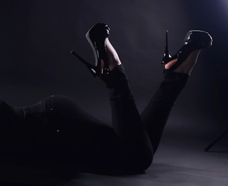 A person is lying down with their legs elevated, wearing glossy black high heels and black pants. The scene is low-lit, creating a dramatic contrast with the dark background.