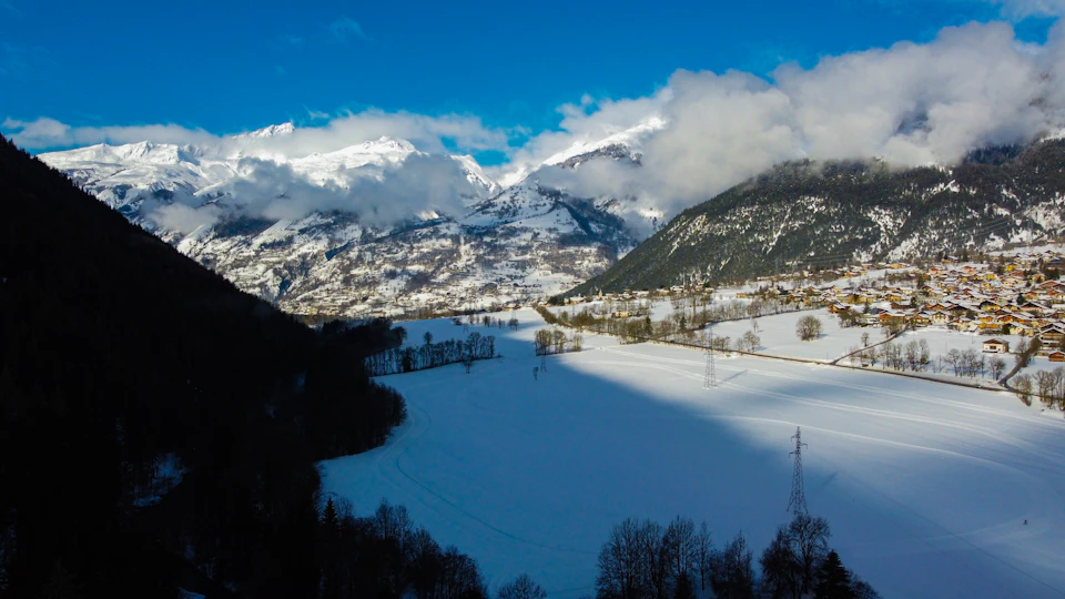 The image presents a breathtaking view of a snowy mountain range under a clear blue sky. The mountains, blanketed in snow, rise majestically against the horizon, their peaks touching the sky. The landscape is dotted with trees and bushes, adding a touch of green to the otherwise white surroundings. In the foreground, there's a valley that stretches out into the distance, its expanse filled with more trees and bushes. A small town can be seen in the background, nestled among these natural elements.