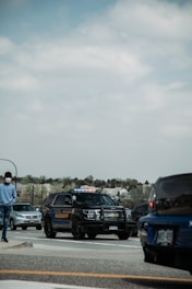 A sheriff's SUV is parked on a road with its lights on. The vehicle is surrounded by other cars, and a person wearing a mask is walking nearby. Trees and houses are visible in the background under a cloudy sky.
