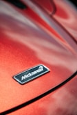 A close-up of a car hood displaying a metallic badge with the brand logo against a glossy, red-orange surface that reflects light.