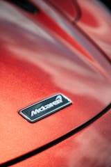 A close-up of a car hood displaying a metallic badge with the brand logo against a glossy, red-orange surface that reflects light.