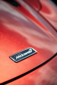 A close-up of a car hood displaying a metallic badge with the brand logo against a glossy, red-orange surface that reflects light.