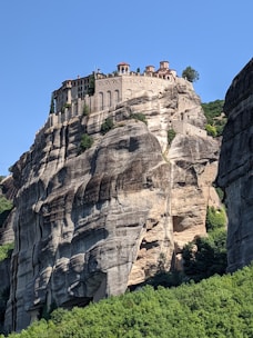 A historic monastery is perched atop a massive cliff formation, surrounded by lush greenery and under a clear blue sky. The structure features arches and red-tiled roofs, indicating its cultural heritage.