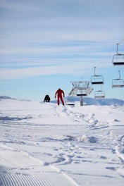 A cheerful family ski sherpa carrying kids' skis and boots from a snowy parking lot toward the ski school lodge.